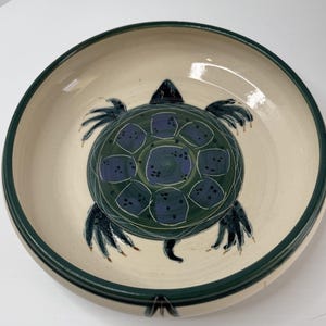 May include: A ceramic bowl with a hand-painted turtle design. The turtle is depicted in shades of green and blue, with a cream-colored background and a dark green rim. The bowl is round and appears to be handmade.