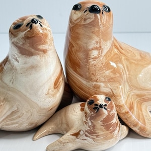May include: Three ceramic seal figurines featuring a marbled pattern of tan, cream, and brown swirls. Each seal has a smooth, glossy finish with painted black eyes and a black nose. The figures are arranged in a group with two larger seals.