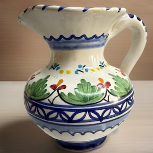 May include: A white ceramic pitcher with a spout and handle, adorned with blue and green floral patterns. The pitcher features a wavy blue border around the top and base, and measures approximately 8 inches in height.