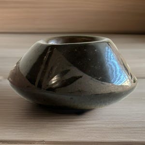 May include: A small, dark brown ceramic bowl with a wide base and a narrow opening. The bowl has a glossy finish and a slightly textured surface. The design features subtle, geometric patterns. The bowl is suitable for holding small items.