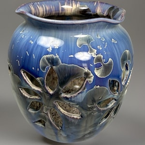 May include: A blue ceramic vase with a unique design. The vase has a round body with cut-out leaf-like shapes and a ruffled rim. The color transitions from a deep blue to lighter shades, with white and gray accents. The vase is approximately 10 inches tall.