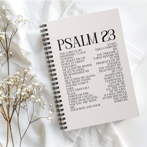 Op de afbeelding: Een spiraalgebonden notitieboek met de tekst "PSALM 23" in grote, vette letters. De pagina's bevatten de volledige tekst van Psalm 23 in zwarte letters. Het notitieboek ligt op een witte ondergrond met delicate witte bloemen.