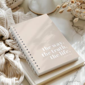 Puede incluir: Un cuaderno con espiral de color beige claro con el texto plateado "the way, the truth, the life." El cuaderno está apilado sobre un libro beige, con una manta de punto color crema en el fondo. El texto "JOHN 14:6" es visible.