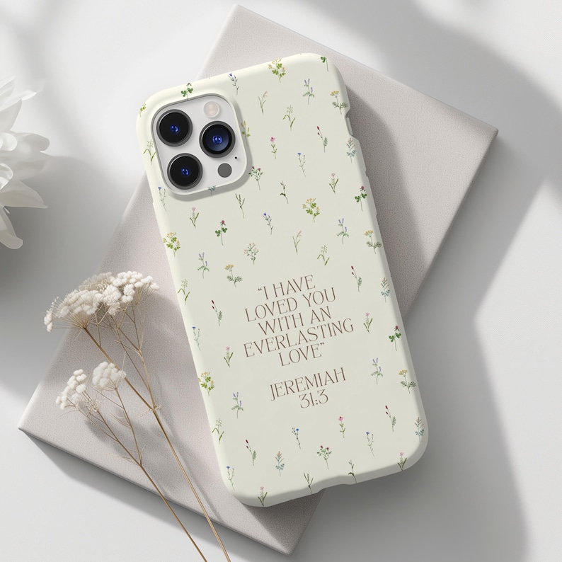 May include: A cream phone case adorned with a delicate floral design and the quote "I HAVE LOVED YOU WITH AN EVERLASTING LOVE" from Jeremiah 31:3. The case is decorated with tiny green, yellow, and purple flowers.