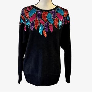 Pode incluir: Camisola preta com decote redondo e mangas compridas. Os ombros e a parte superior do peito são adornados com um design de lantejoulas coloridas em tons de vermelho, laranja, rosa, roxo e turquesa. A camisola tem uma textura canelada.