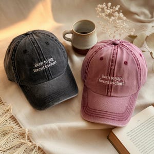 May include: Two vintage-style baseball caps with the text "Born to yap forced to chart" embroidered on the front. One cap is dark gray and the other is a dusty rose color.