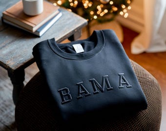 May include: A dark gray sweatshirt with the word "BAMA" embroidered in a lighter gray font. The sweatshirt is folded and resting on a patterned stool. A small wooden table with books and a mug is in the background.