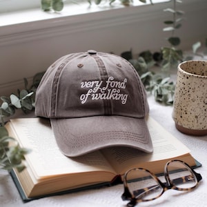 May include: A brown baseball cap with the embroidered text "very fond of walking" in white. The cap is resting on an open book, with a pair of glasses and a speckled mug nearby. The scene is set near a window with greenery.