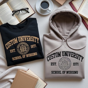 Peut inclure: Un sweat-shirt noir et un sweat à capuche beige, brodés avec "CUSTOM UNIVERSITY EST. 1971 SCHOOL OF NURSING". L'image présente les vêtements avec des livres, des lunettes et une tasse de café.