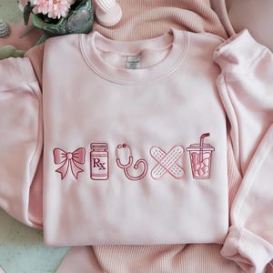 May include: A light pink sweatshirt with embroidered medical-themed designs. The designs include a bow, prescription bottle, stethoscope, crossed bandages, and a drink with a straw. The sweatshirt is made of a soft material.