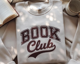 Puede incluir: Sudadera beige con las palabras "BOOK Club" en una fuente granate de estilo universitario. La sudadera está doblada cuidadosamente, con las mangas y el cuello visibles. Un libro, gafas y una taza están en el fondo.