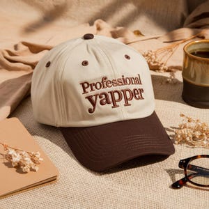 May include: A beige baseball cap with a brown brim and the embroidered text "Professional Yapper" in brown. The cap has a brown button on top and brown eyelets. The hat is on a textured surface.