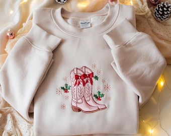 Peut inclure: Un sweat-shirt crème avec un motif brodé de bottes de cowboy roses avec des nœuds rouges, du houx et des flocons de neige. Ce haut à manches longues est un vêtement confortable et festif, parfait pour la saison des fêtes.