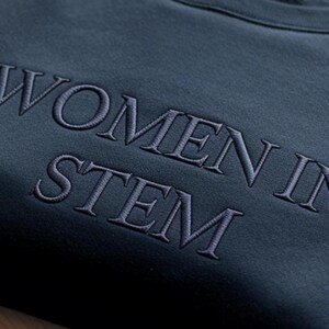 Może przedstawiać: Ciemnoniebieska bluza z haftowanym napisem "WOMEN IN STEM" w jaśniejszym odcieniu niebieskiego. Bluza jest złożona, eksponując wyraźnie napis.