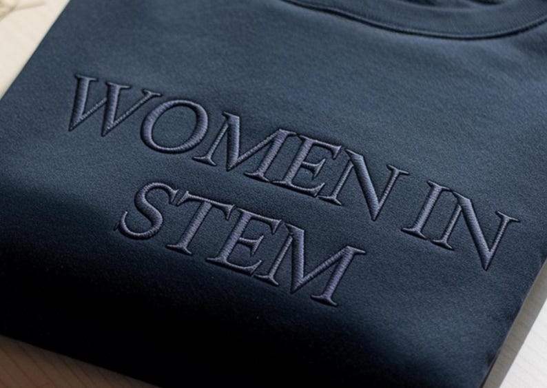 Może przedstawiać: Ciemnoniebieska bluza z haftowanym napisem "WOMEN IN STEM" w jaśniejszym odcieniu niebieskiego. Bluza jest złożona, eksponując wyraźnie napis.