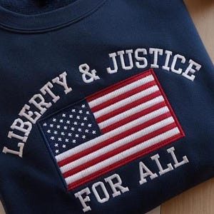 May include: Navy blue sweatshirt with the embroidered words "LIBERTY & JUSTICE FOR ALL" in white. The design includes an embroidered American flag in red, white, and blue.