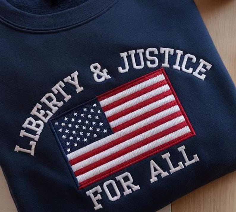 May include: Navy blue sweatshirt with the embroidered words "LIBERTY & JUSTICE FOR ALL" in white. The design includes an embroidered American flag in red, white, and blue.