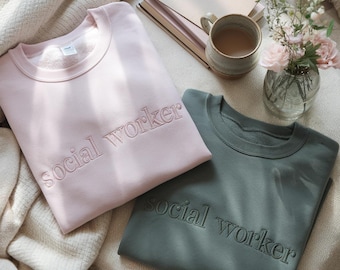 Könnte beinhalten: Zwei Sweatshirts, eines rosa und eines grün, mit dem Text "social worker" bestickt.
