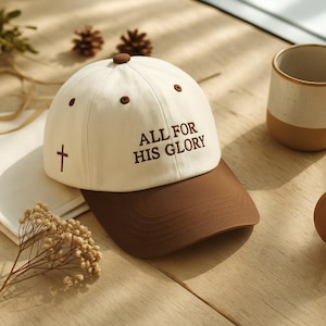 May include: A baseball cap with a white crown and a brown peak. The cap has the embroidered text "ALL FOR HIS GLORY" and a cross symbol. The cap is on a wooden surface.