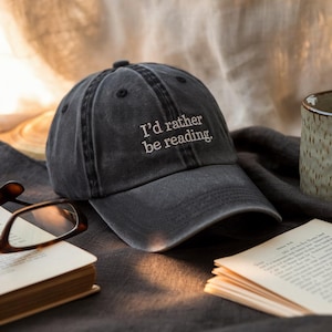 May include: A dark gray baseball cap with the embroidered text "I'd rather be reading." The hat is sitting on a dark gray surface next to a book and a pair of glasses. A ceramic mug is in the background.