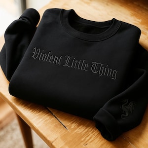 May include: Black sweatshirt featuring the embroidered text "Violent Little Thing" in a gothic style. A dragon design is on the sleeve. The garment is folded on a wooden surface.