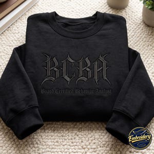 May include: A black long-sleeve sweatshirt featuring dark grey embroidery on the chest. The design includes the large gothic-style letters BCBA with the text Board Certified Behavior Analyst embroidered in a smaller font directly underneath.