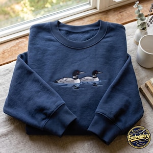 May include: A navy blue crewneck sweatshirt featuring an embroidered design of two loons swimming on water. The birds have black heads, red eyes, and white and grey patterned wings. The sweatshirt has long sleeves with ribbed cuffs and a ribbed collar.