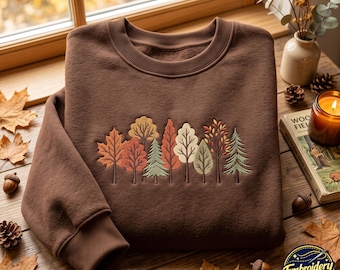 May include: A dark brown crewneck sweatshirt featuring an embroidered design of a forest line with various trees in shades of orange, yellow, green, and cream. The sweatshirt has ribbed cuffs and a ribbed neckline, showcasing a detailed nature motif.
