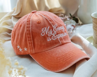 May include: An orange baseball cap with the embroidered text "My Energy is Expensive" in white script. Three white star designs are embroidered on the side. The hat has a curved brim and a fabric strap.