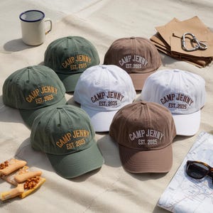 May include: Several baseball caps in shades of green, white, and brown. Each cap displays the embroidered text "CAMP JENNY" and the year "EST. 2023" or "EST. 2025". The caps are arranged on a light-colored, textured surface.