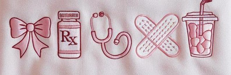 May include: Pink embroidered design featuring a bow, prescription bottle with "Rx" text, stethoscope, crossed bandages, and a drink with a straw. The design is on a white background.