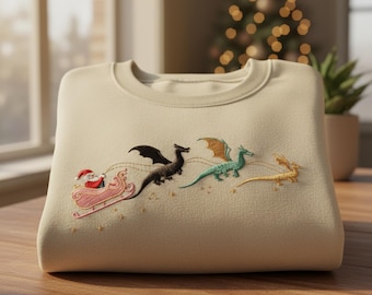 May include: A beige sweatshirt featuring an embroidered design of Santa Claus in a pink sleigh pulled by three dragons. The dragons are black, green, and gold. The design is set against a background of small gold stars.