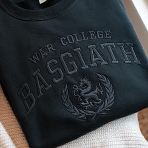May include: Black sweatshirt featuring embroidered text: "WAR COLLEGE BASGIATH". The design includes an embroidered dragon emblem within a laurel wreath. The garment is made from a soft fabric, suitable for casual wear.