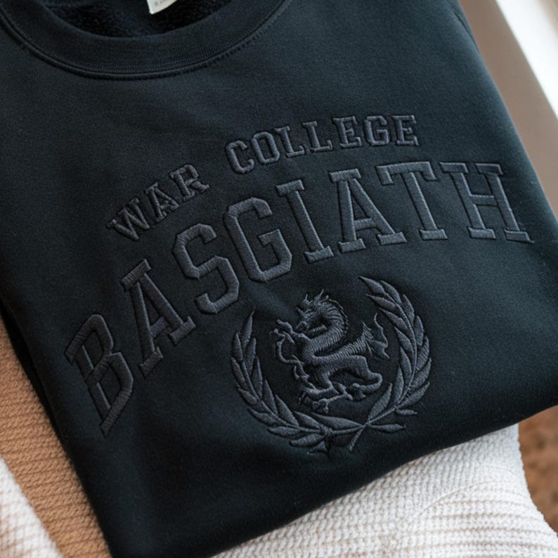 May include: Black sweatshirt featuring embroidered text: "WAR COLLEGE BASGIATH". The design includes an embroidered dragon emblem within a laurel wreath. The garment is made from a soft fabric, suitable for casual wear.