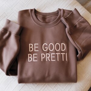 May include: A brown sweatshirt with the embroidered text "BE GOOD BE PRETTI" in white. The sweatshirt has a crew neck and long sleeves, folded neatly on a white surface. The fabric appears soft and comfortable.