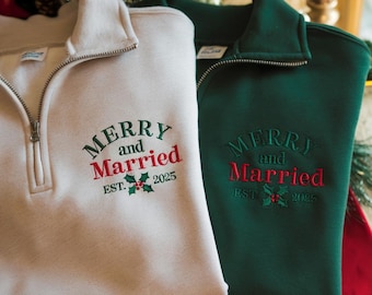 Può includere: Due felpe con zip a un quarto, una color crema e una verde scuro. Entrambe presentano la scritta ricamata "MERRY and Married EST. 2025" in verde e rosso, con accenti di agrifoglio. Le felpe hanno un collo e una cerniera argentata.