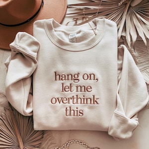 Puede incluir: Sudadera color crema con la frase bordada "hang on, let me overthink this" en fuente marrón. La parte superior de manga larga está doblada, con un sombrero y elementos decorativos en el fondo.