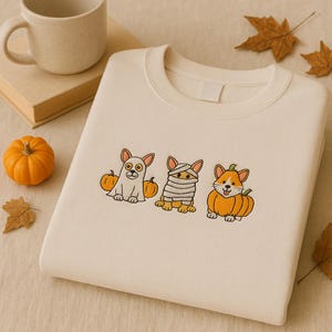 Może przedstawiać: Kremowa bluza z haftowanymi corgi w tematyce Halloween. Jeden corgi jest przebrany za ducha, drugi za mumię, a trzeci siedzi w dyni. Jesienne liście i mała dynia otaczają bluzę.