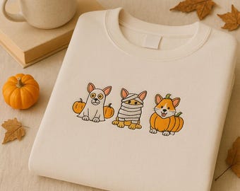 May include: Cream-colored sweatshirt featuring embroidered Halloween-themed corgi dogs. One corgi is dressed as a ghost, another as a mummy, and the third is in a pumpkin. A small orange pumpkin and fall leaves are also visible.