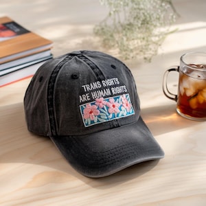 Può includere: Un cappello da baseball in denim nero con il testo "Trans Rights Are Human Rights" ricamato in bianco sul davanti. Il testo è circondato da un rettangolo blu e verde con fiori rosa ricamati.