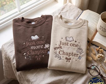 May include: Two crewneck sweatshirts, one dark brown and one beige, each featuring embroidered text that reads Just one more Chapter. The design includes icons of an open book, eyeglasses, a coffee mug, a small floral sprig, and a bow.