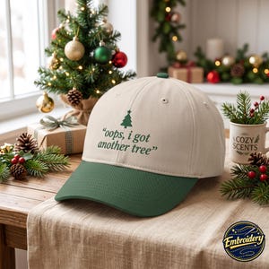 Peut inclure: Une casquette de baseball beige avec une visière vert foncé. L'avant présente une petite icône de sapin de Noël vert au-dessus du texte vert foncé qui indique "oops, i got another tree" dans une police simple en minuscules.