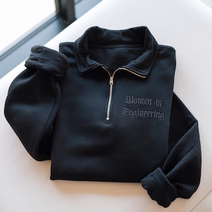 May include: Black sweatshirt with a collared neckline and a gold zip. The text "Women in Engineering" is embroidered on the front. The sleeves are cuffed. The sweatshirt is folded on a white surface.
