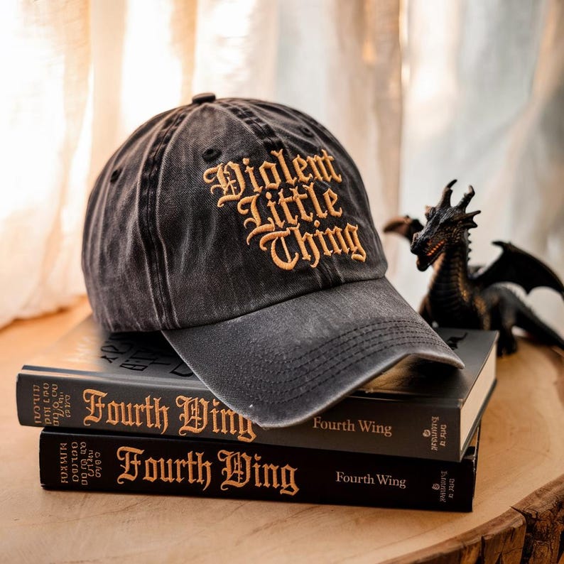 May include: A black baseball cap with a gold embroidered design that reads "Violent Little Thing". The cap is sitting on top of two books with the title "Fourth Wing" on the cover.