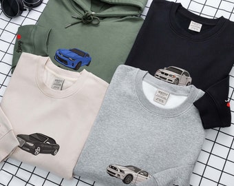 May include: Five different sweatshirts with various car designs embroidered on them. The sweatshirts are in different colours, including beige, grey, black, and green. The car designs include a blue sports car, a black saloon, a white saloon, and a grey muscle car.