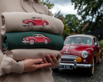 May include: Three folded hoodies in beige and dark green, each featuring an embroidered red vintage car. The hoodies are held by a person, with a matching red classic car visible in the background.