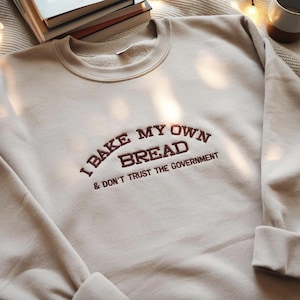 Op de afbeelding: Beige sweatshirt met de geborduurde tekst "I BAKE MY OWN BREAD & DON'T TRUST THE GOVERNMENT" in bruin. De sweatshirt heeft een ronde hals en lange mouwen met geribde manchetten. De stof ziet er zacht en comfortabel uit.