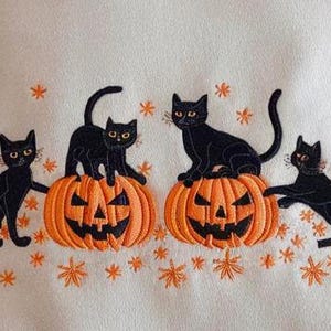 May include: Embroidered design featuring four black cats with yellow eyes, posed around two orange jack-o'-lanterns. The design includes orange star-shaped accents on a cream-colored background, evoking a Halloween theme.