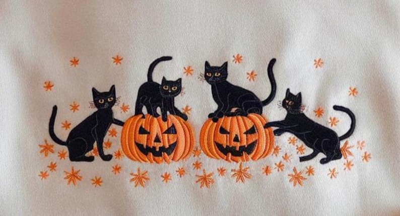May include: Embroidered design featuring four black cats with yellow eyes, posed around two orange jack-o'-lanterns. The design includes orange star-shaped accents on a cream-colored background, evoking a Halloween theme.
