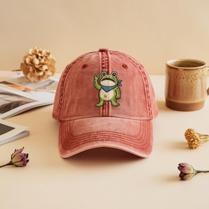 May include: A faded red baseball cap with an embroidered frog motif. The frog wears a blue bandana. A ceramic mug and dried flowers are in the background. The cap has a distressed finish.
